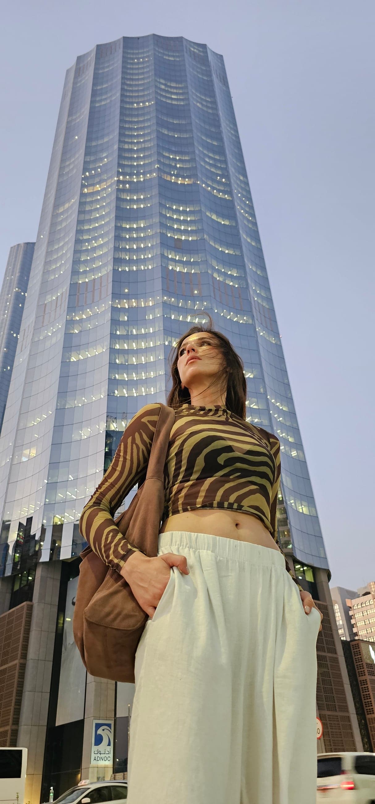 Olga Kovaleva in a zebra-print mesh top and white linen trousers in front of a glass skyscraper at dusk